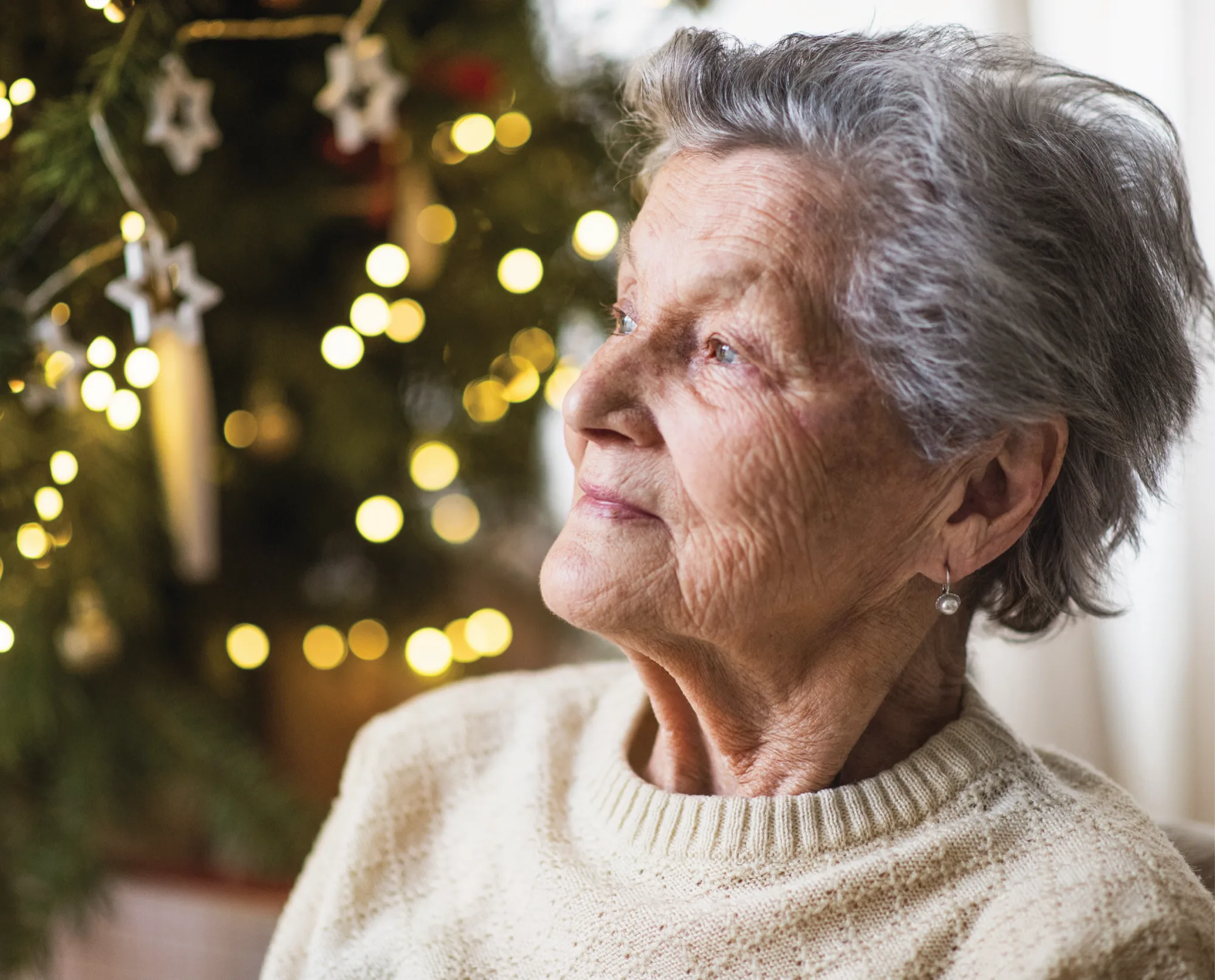 An older woman in a cream jumper gazing out of a window, with a Christmas tree and twinkling lights behind her