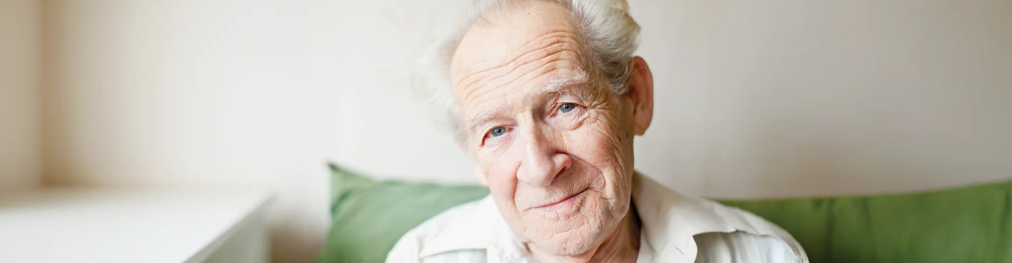An older man with white hair smiling gently while sitting on a sofa with green cushions