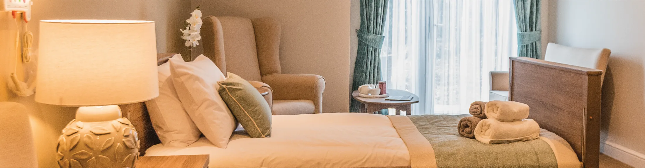 A care home bedroom at Colwall with a made-up bed, lamp, armchairs and a tea tray by the window