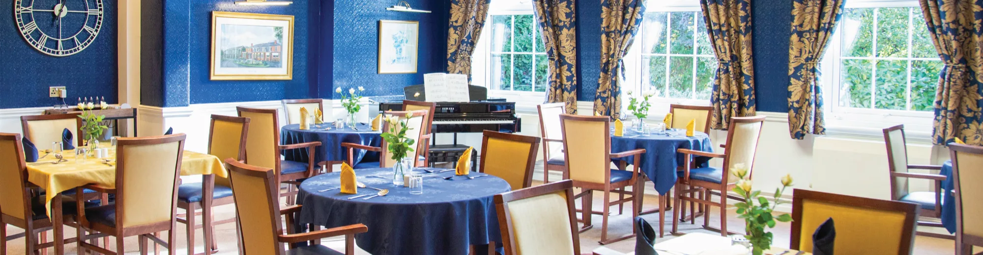 The dining room at Dorset House, with blue and gold laid tables, a piano and tall windows framed by patterned curtains