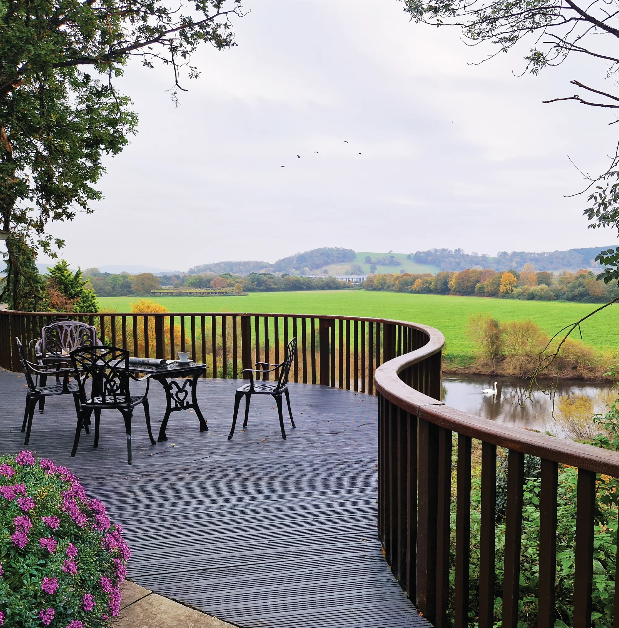 A curved wooden terrace with garden furniture overlooking open fields and a river