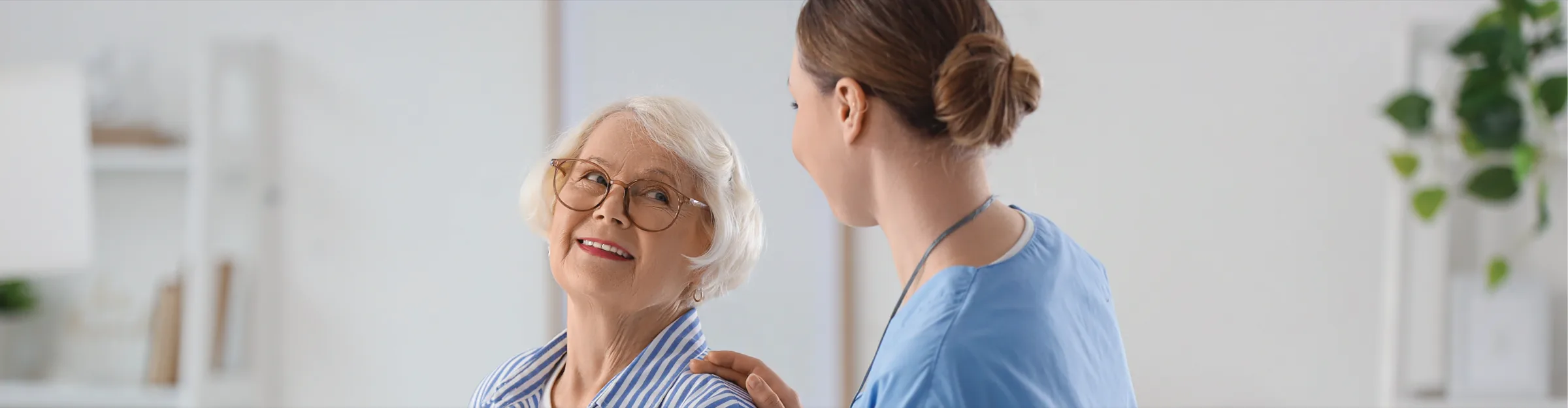 A smiling older woman in glasses looking up at a nurse in blue scrubs who has a hand on her shoulder