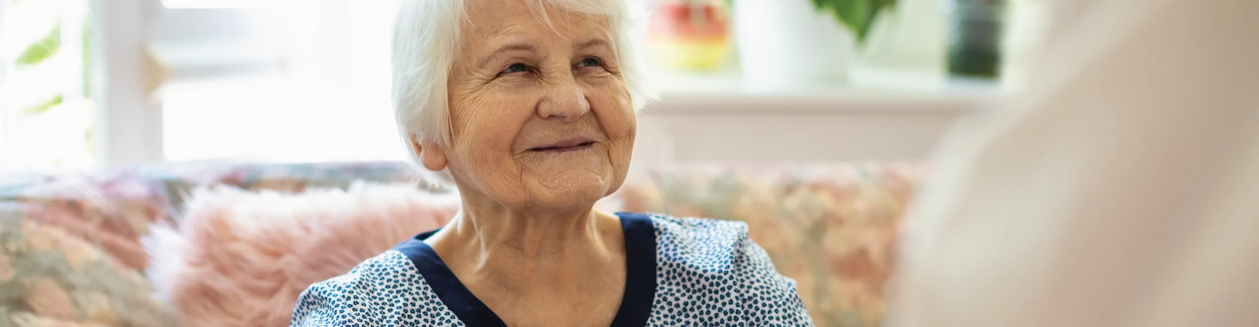 An older woman with white hair smiling as she sits on a floral sofa beside a sunlit window