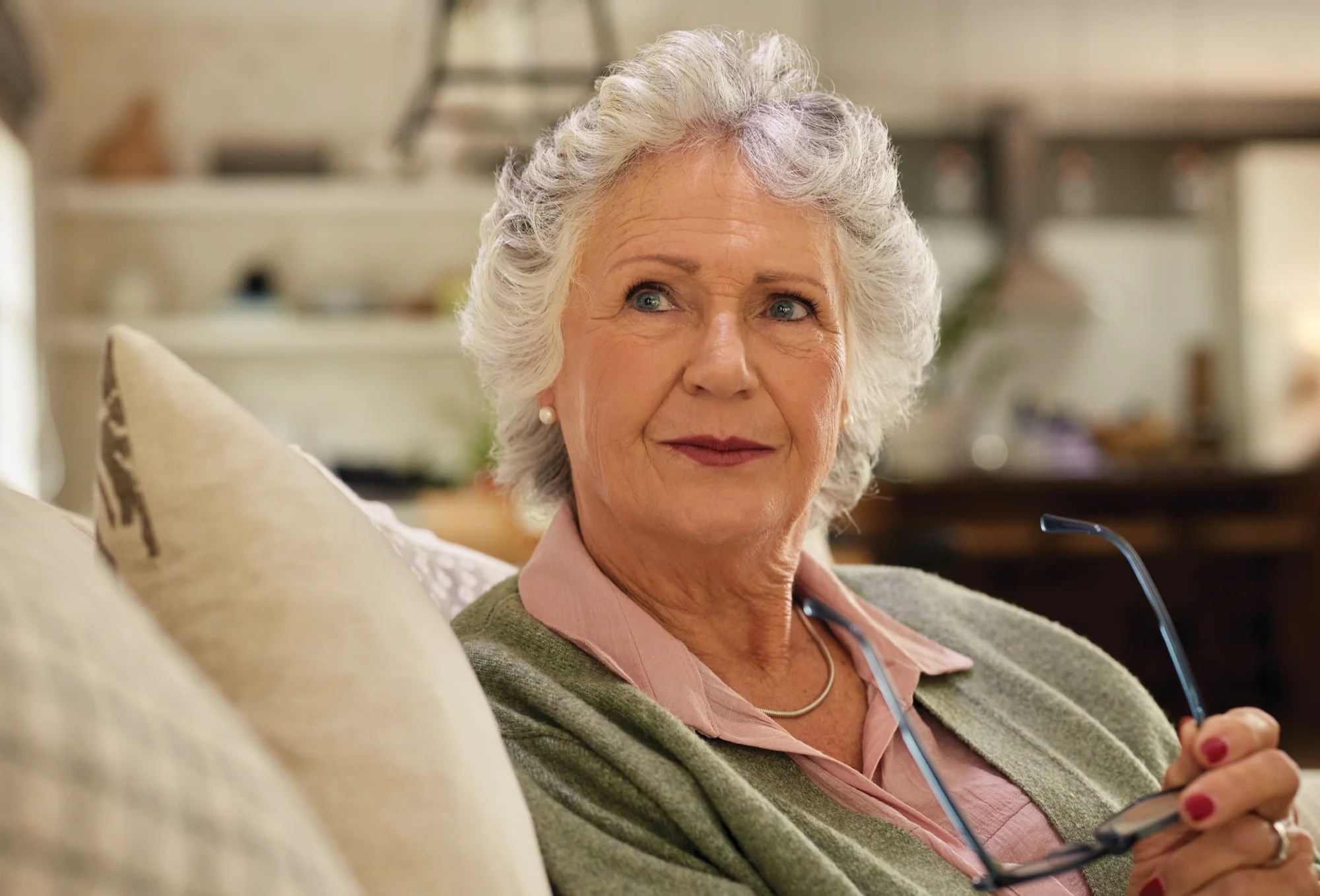 An older woman sitting on a sofa holding her glasses, looking thoughtfully into the distance