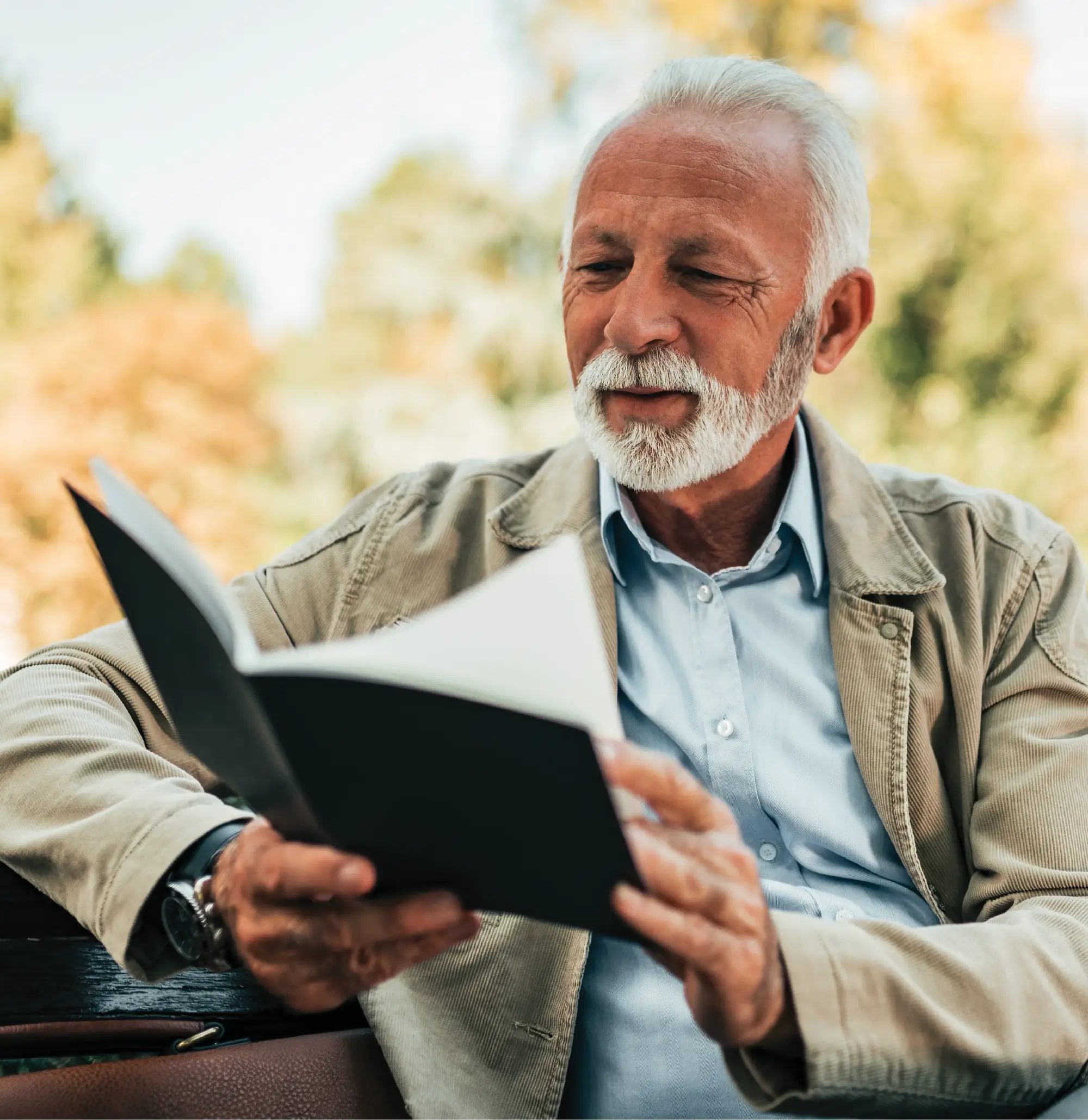 An older man with a white beard reading a book outdoors on an autumn afternoon