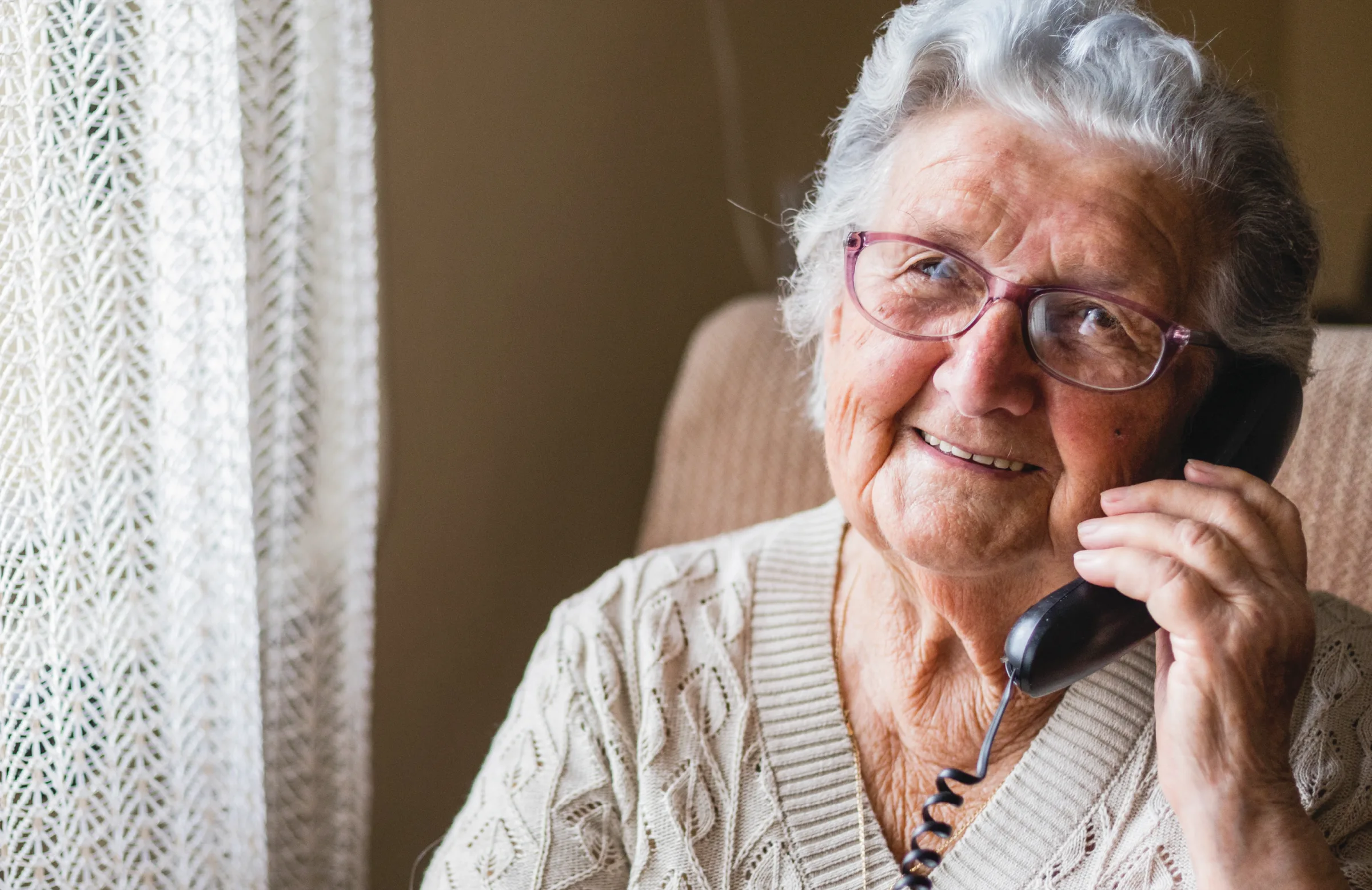 A smiling older woman in glasses talking on a corded telephone beside a lace-curtained window