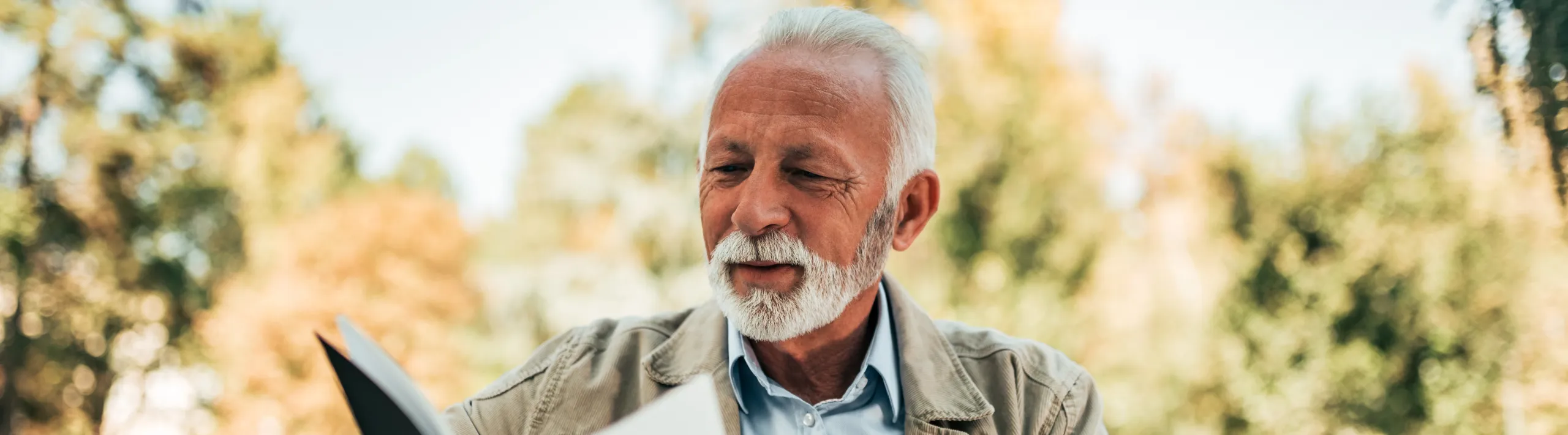 An elderly man reading a book outdoors in autumn sunshine