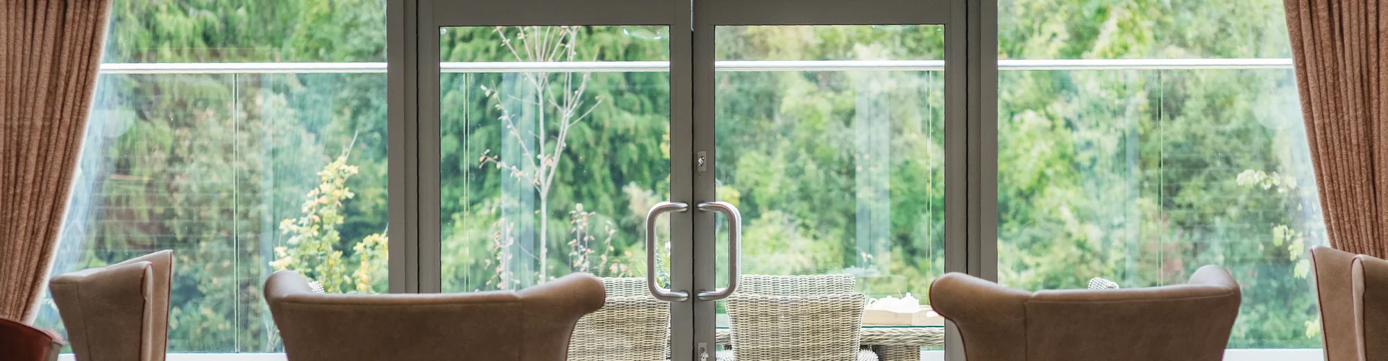 Care home lounge armchairs facing patio doors that open onto a leafy garden terrace