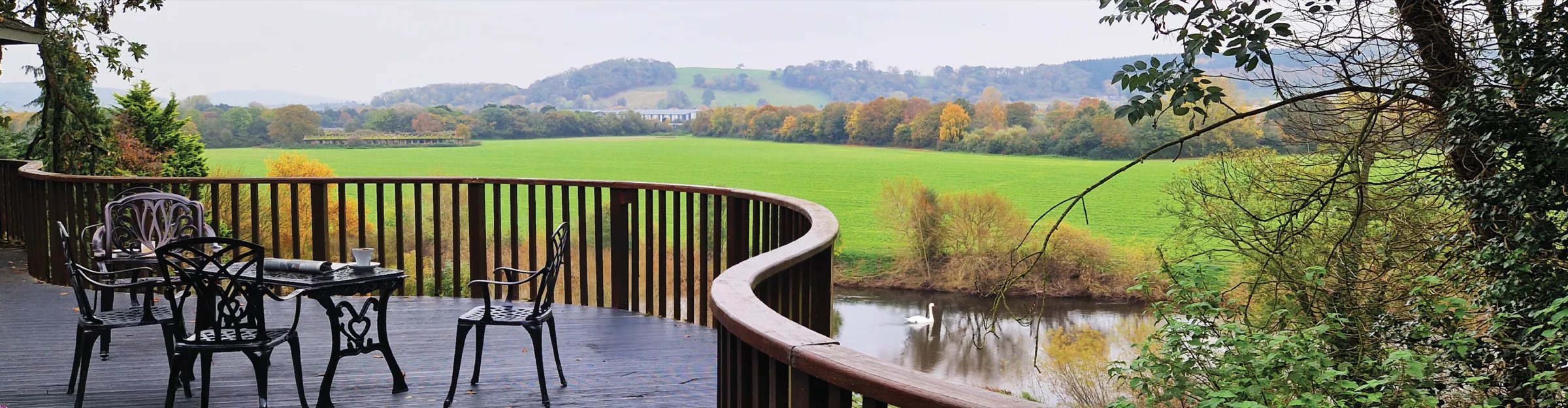 Garden furniture on a curved timber deck at Hampton Gwen, overlooking open fields and a river with a swan