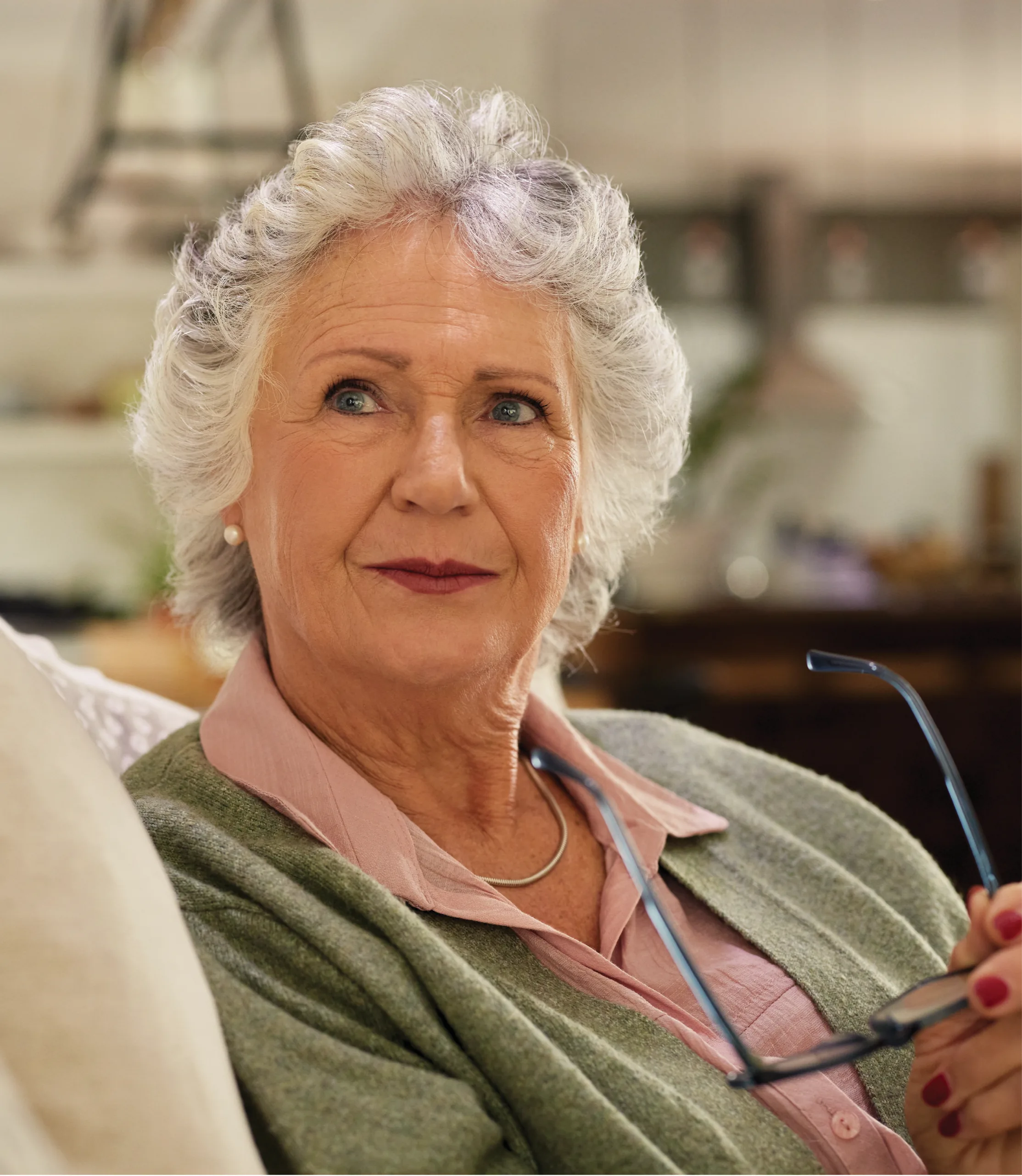 An older woman in a green cardigan holding her glasses and looking thoughtfully into the distance