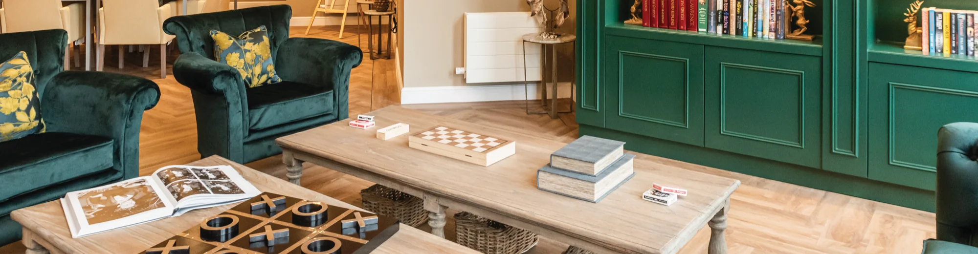 A Colwall care home lounge with green velvet armchairs, a chess set on a coffee table and a green bookcase