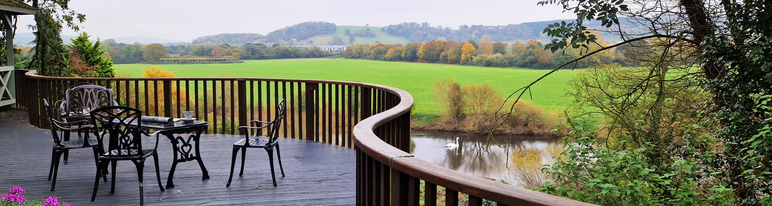 A curved wooden terrace with garden furniture overlooking open fields and a river with a swan