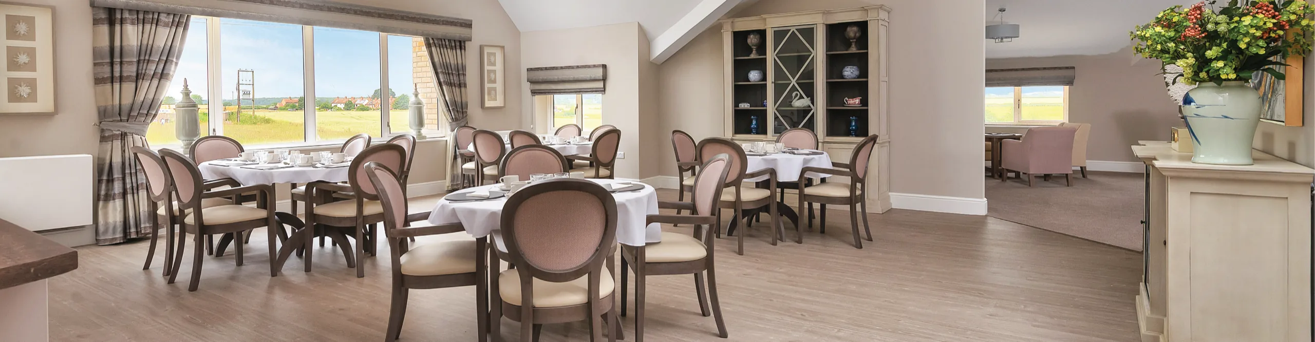 A care home dining room with round tables laid with white linen and countryside views through the windows