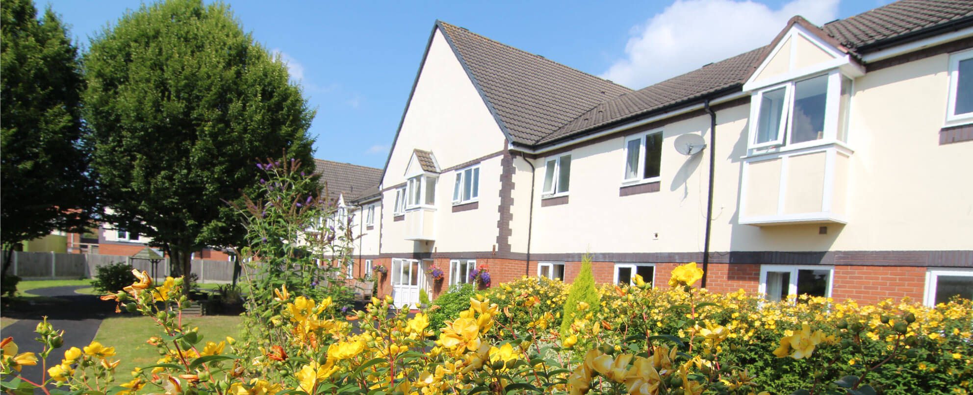 A two-story white building with a brown roof, surrounded by green bushes and yellow flowers under a clear blue sky