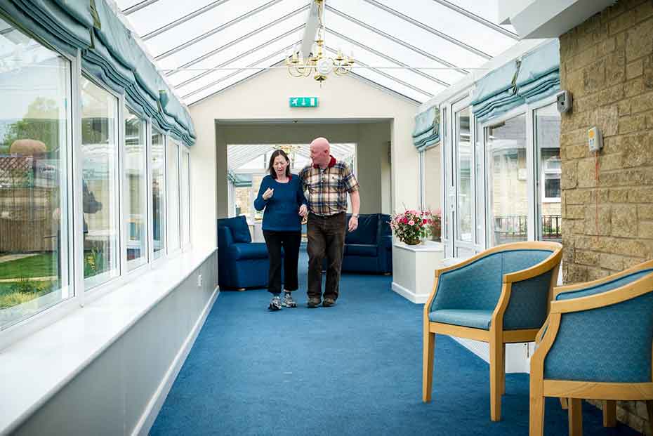 A lady and a man walking in a well-lit corridor with big windows, blue carpet, and chairs