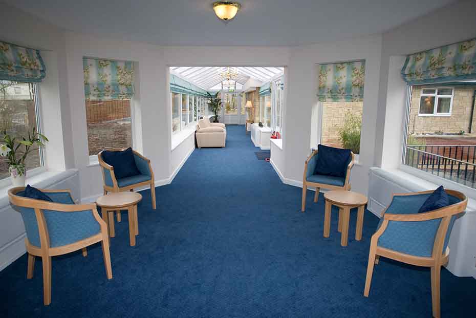 A spacious corridor with blue carpet, white walls, multiple chairs with blue cushions, and large windows allowing ample natural light