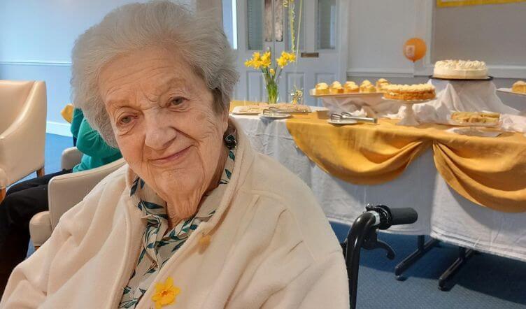 An elderly lady seated indoors, near a table with cakes and refreshments in the background