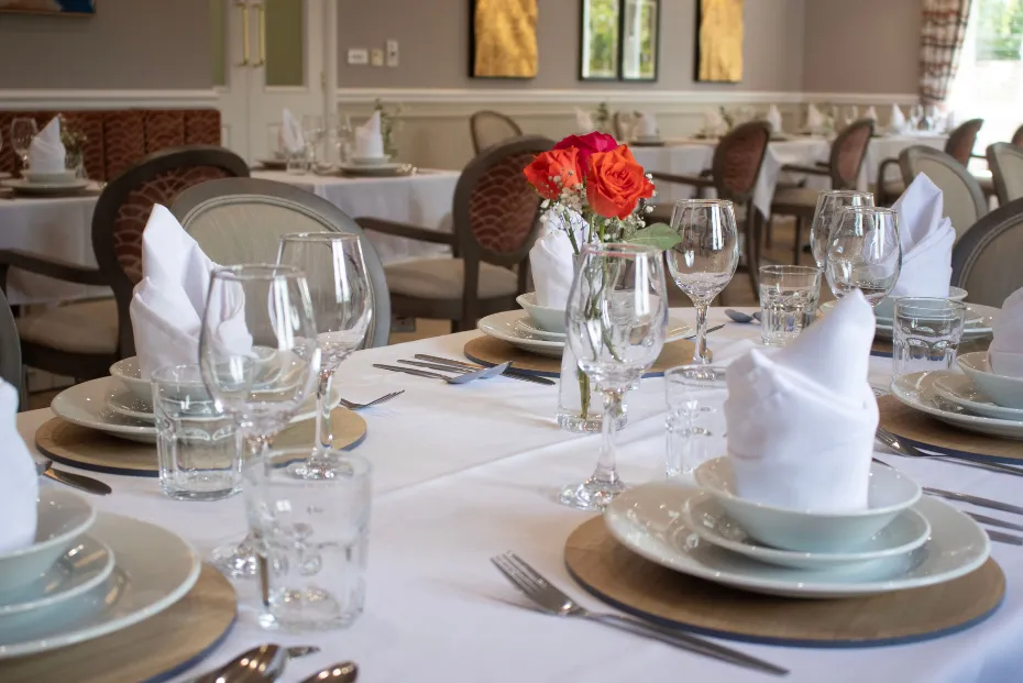 An elegantly set dining table with white linen, folded napkins, crystal glassware and a vase of orange roses, with further set tables in the bright dining room behind
