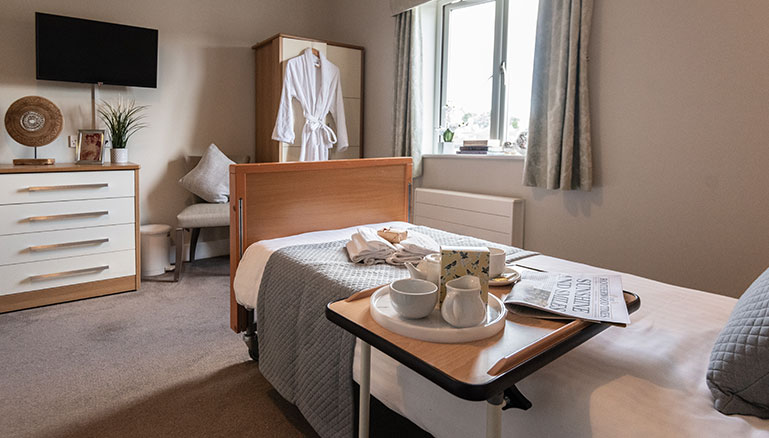 A cosy bedroom interior with natural lighting featuring a single bed adorned with gray bedding next to an overbed table set for tea or coffee service and newspapers spread out; across from which is mounted TV above dresser, beside an armchair and round side table near wardrobe with hanging white robe.