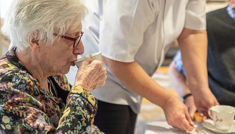 An elderly lady wearing a colourful floral shirt with white hair, is seated at a table drinking a cup of tea.