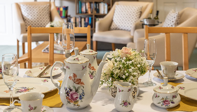 A well-arranged dining table set for tea. On the table there are porcelain teapots with floral patterns next to a matching creamer and a bouquet of white flowers in a small vase. Surrounding these items are several teacups and saucers with similar designs, along with clear glasses.