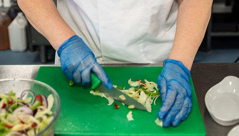 A person wearing a white chef’s uniform and blue gloves is chopping vegetables on a green cutting board. There is a bowl of sliced vegetables to the left.