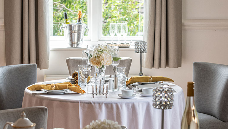 An elegantly set round dining table in a bright room with windows. The table is adorned with a white tablecloth and set with silver cutlery, crystal glassware, a bouquet of white flowers, and a champagne bucket with a bottle. Two grey upholstered chairs are positioned at the table.