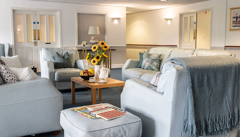 A cosy living room setting with two light grey sofas adorned with various patterned cushions. A wooden coffee table between the sofas holds a bowl of fruit, a vase with sunflowers, and some magazines. 