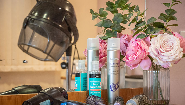 A countertop featuring a black salon hair dryer helmet, hairspray, hairbrushes and combs, and a vase with pink roses.