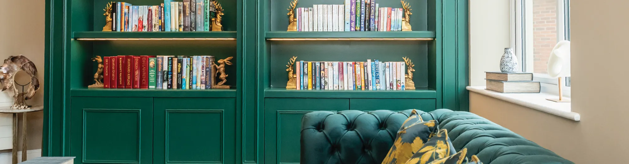 Green built-in bookshelves lined with books and gold stag bookends, beside a green velvet sofa with patterned cushions