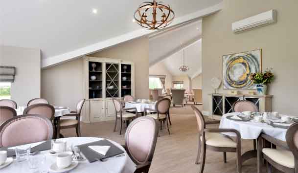 An elegant dining room with round tables set for a meal, pink upholstered chairs, a chandelier, and an abstract painting on the wall