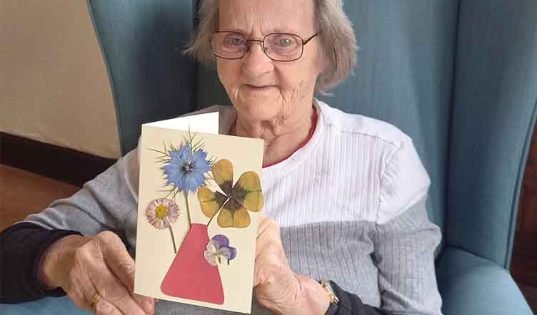 Elderly lady sitting in a blue armchair holding a card with pressed flowers on the front