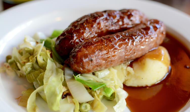 Plate of food featuring two grilled sausages, mashed potatoes with gravy, and cooked cabbage