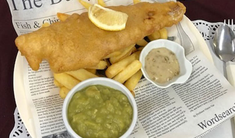 Plate of traditional British fish and chips served with a side of mushy peas and tartar sauce, served on a newspaper