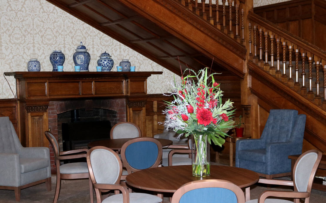 An elegant seating area with a wooden staircase, a fireplace, and a dining area with chairs around a table featuring a vase of red and white flowers