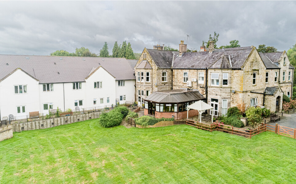 Aerial view of a large traditional stone house with a conservatory extension, adjacent to modern white townhouses, surrounded by a well-kept lawn