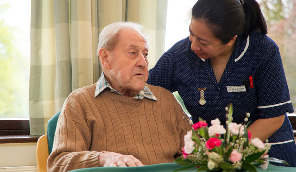 Elderly man is seated at a table with a nurse standing beside them offering support