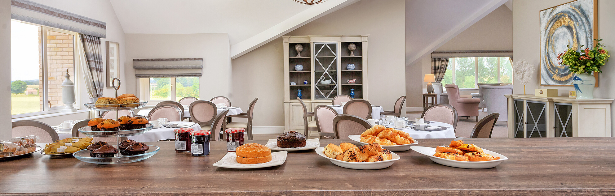 A spacious dining room with a large wooden table set with various pastries and cakes, overlooking a bright window with a view of the outdoors.