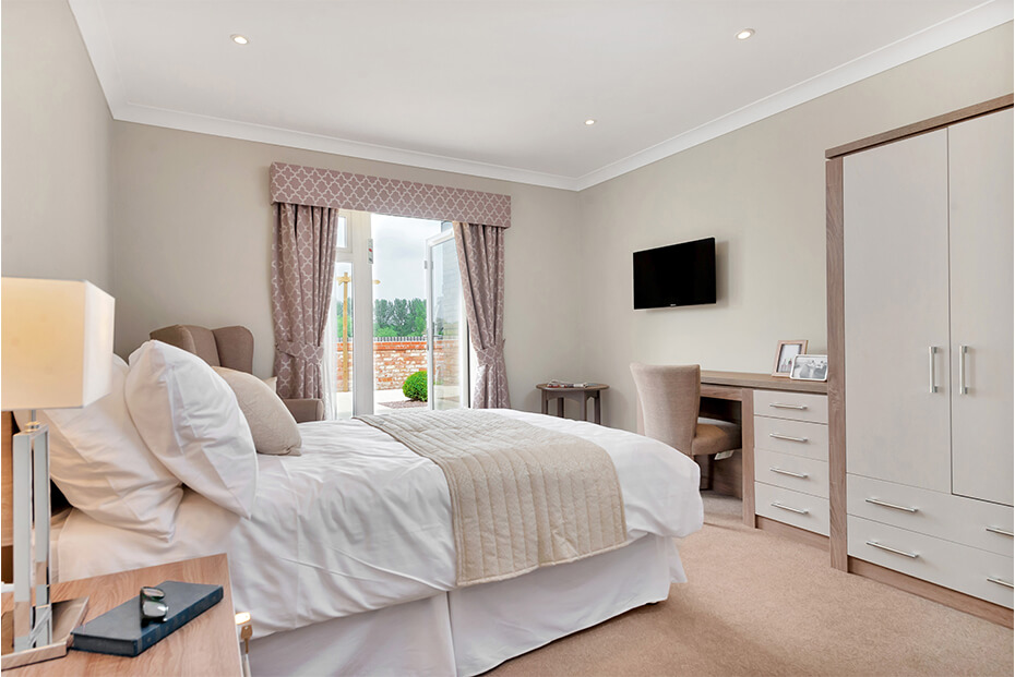 A well-lit, modern bedroom with a large bed, white bedding, a cream throw blanket, two bedside tables with lamps, a desk and chair, and a wall-mounted television.