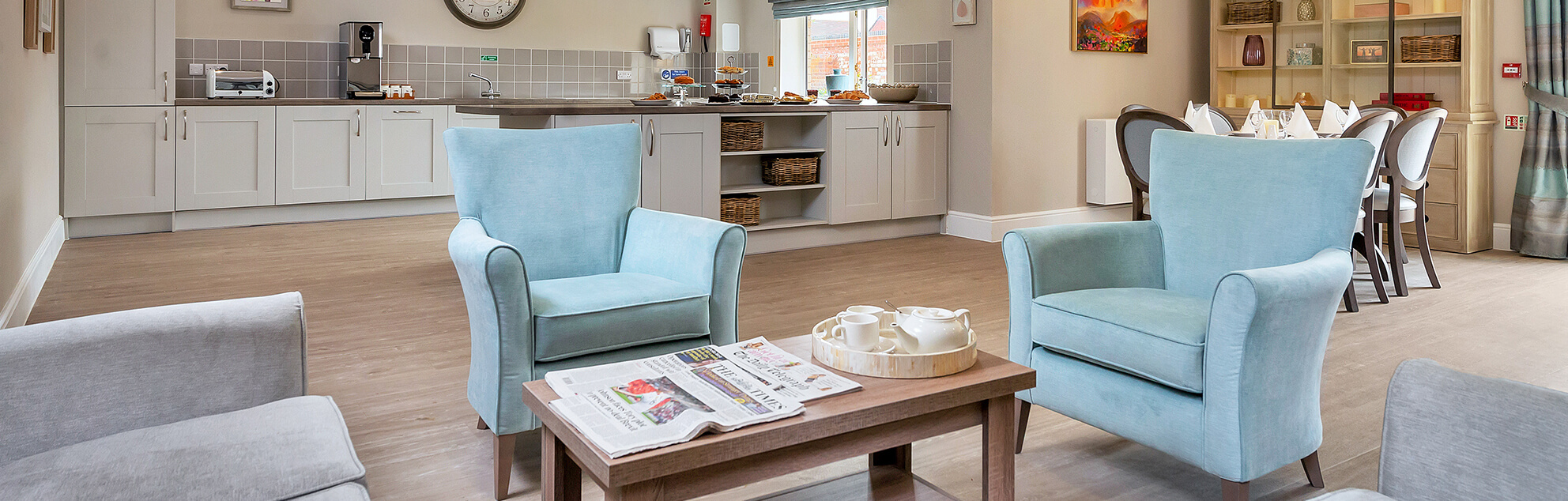 A modern kitchenette and lounge area with two blue armchairs, a wooden coffee table with a teapot set, and newspapers, well-lit with natural light from large windows