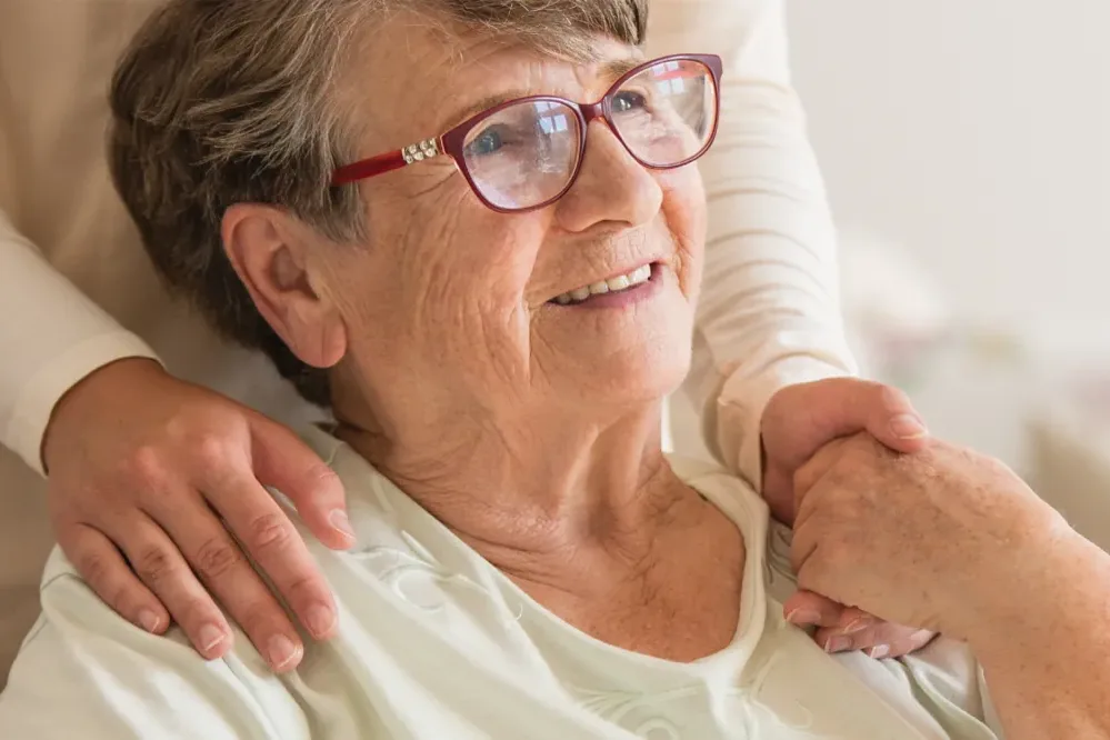 A carer with an arm around the shoulders of a smiling older woman wearing red glasses