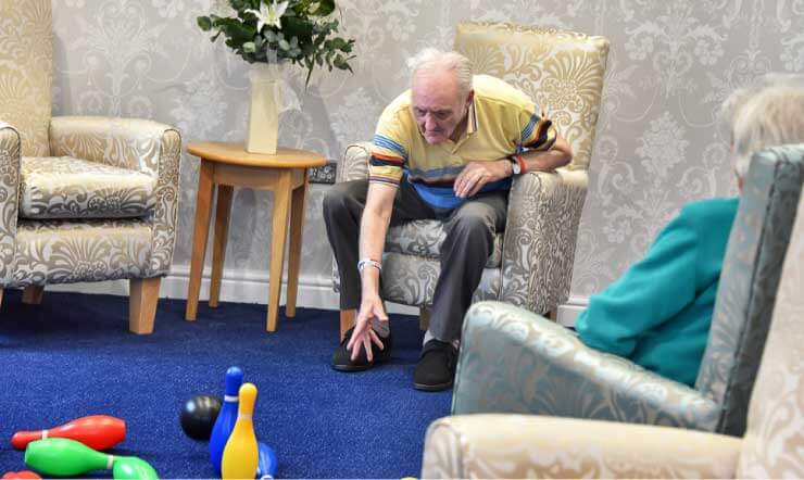 Elderly man seated in a chair, playing a game of skittles on the carpet of a well-furnished living room