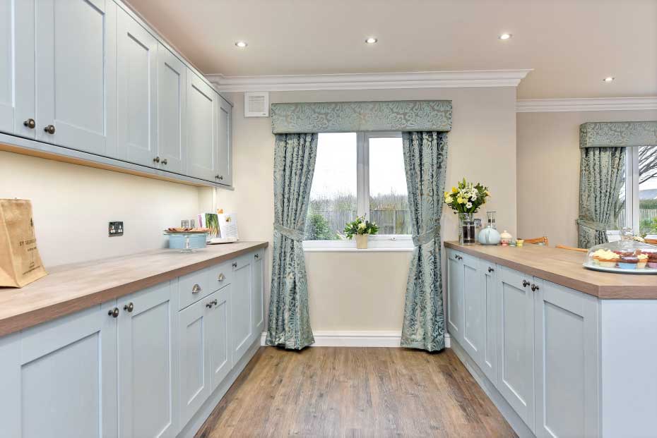 A modern kitchenette with blue cabinets, wooden countertops, a window with patterned curtains, and various kitchen items and decor.