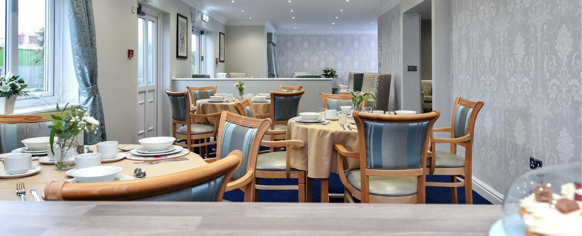 A sophisticated dining room with light grey patterned wallpaper. The room is furnished with multiple round tables set with white plates, silverware, and glassware. Blue upholstered chairs surround the tables.