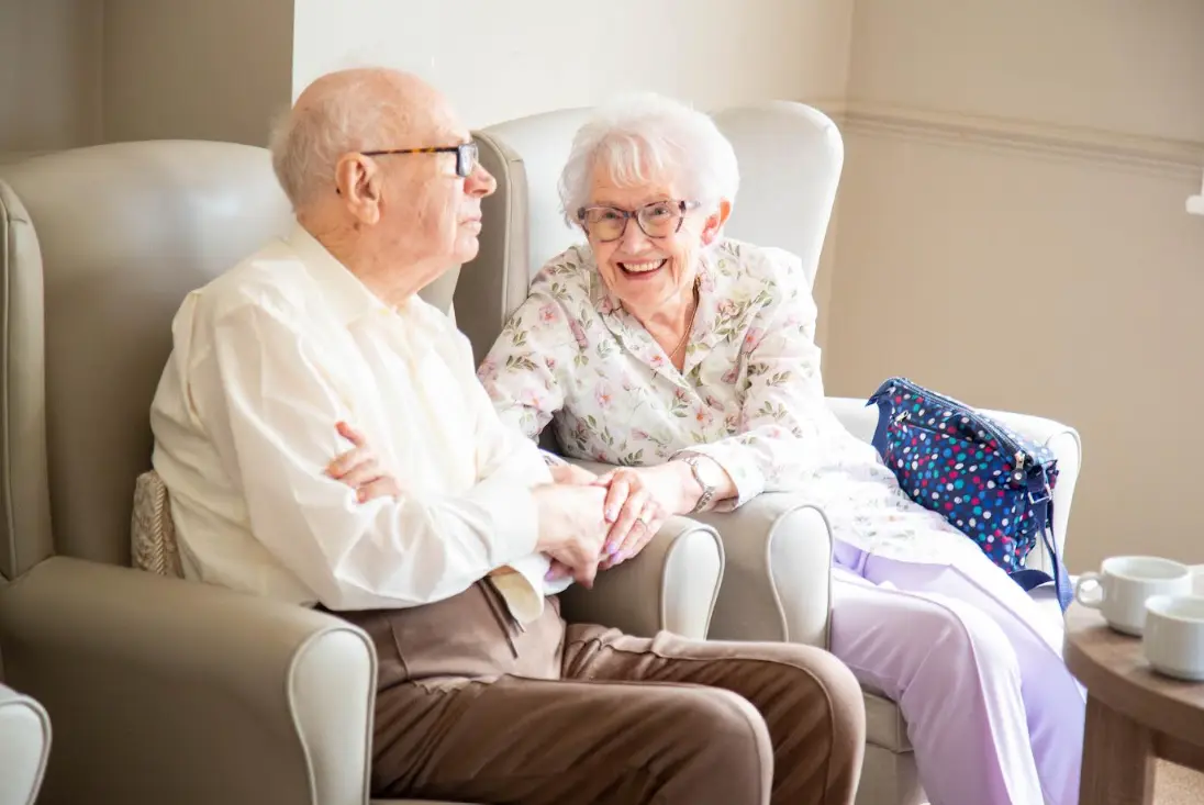 Two Dorset House residents sitting side by side in armchairs, holding hands and smiling
