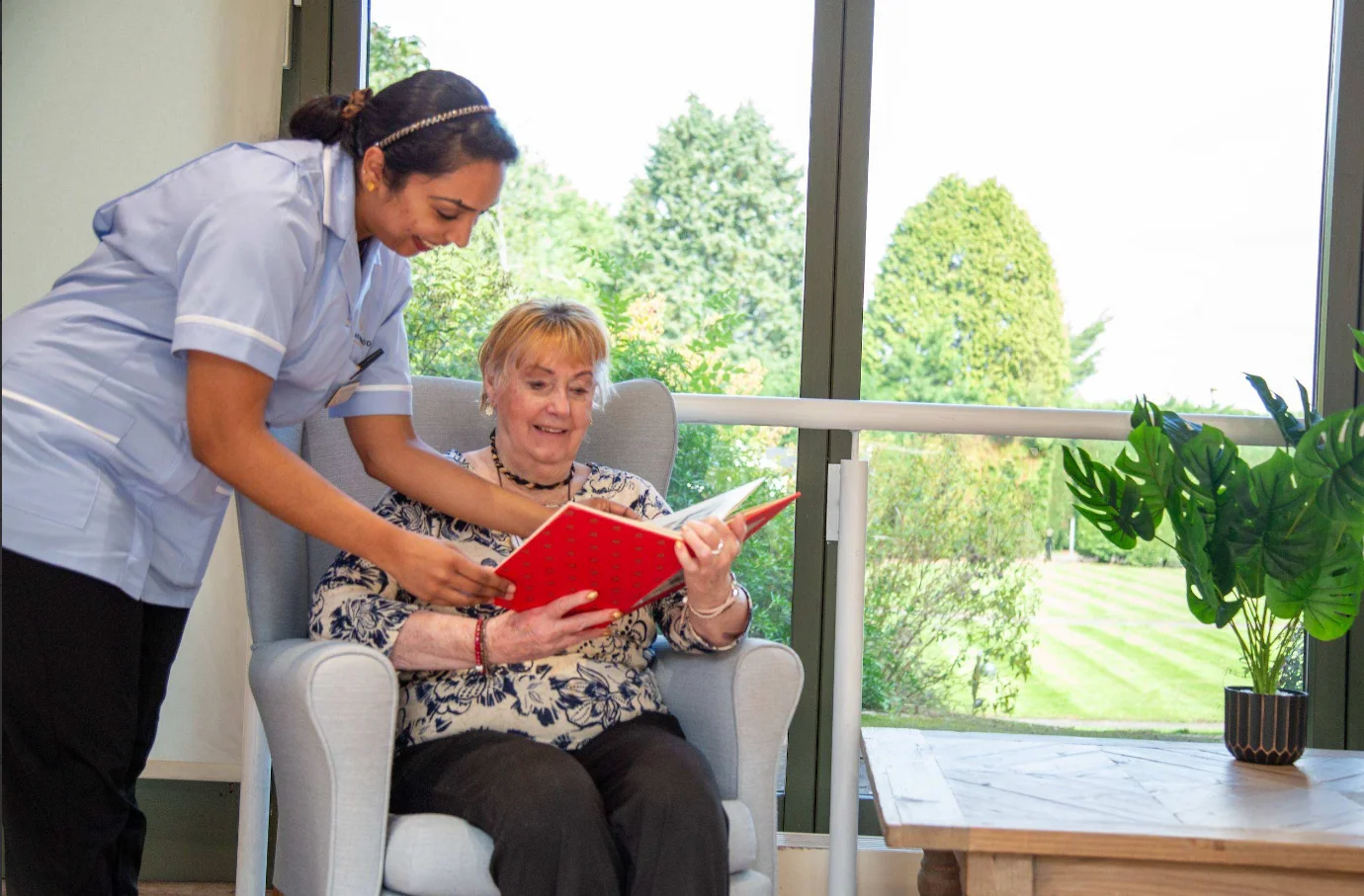 A carer leaning in to look through a book with a resident seated by the garden windows at Dorset House