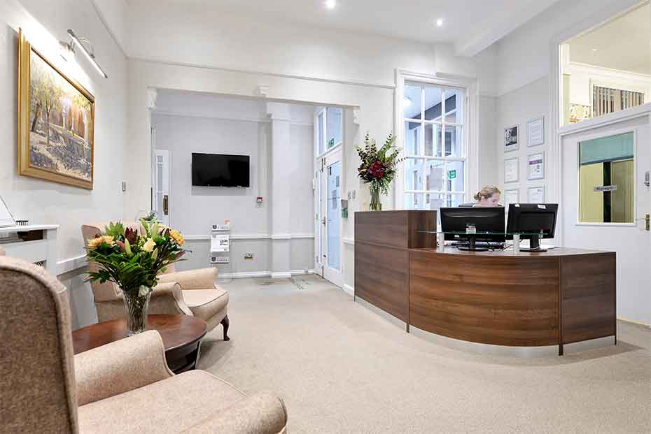 Reception area with a wooden desk with a person working on a computer, a seating area with chairs and a sofa, a wall-mounted TV, and decorative plants.