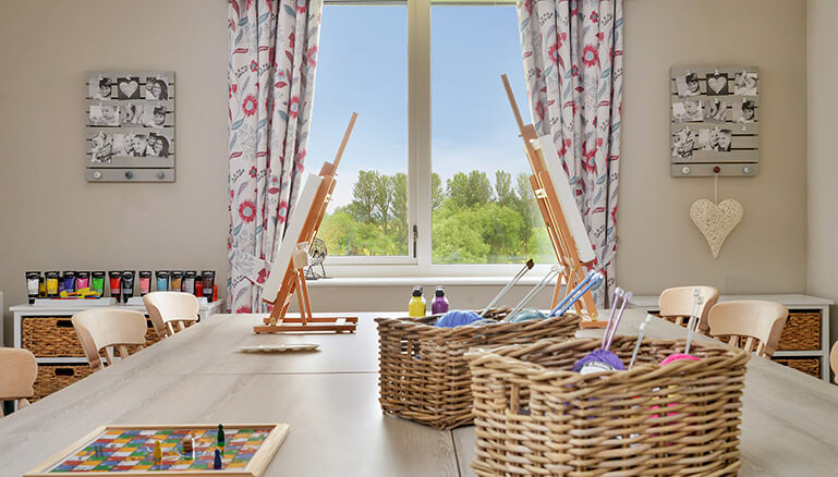 A spacious and organised craftroom with a large table, chairs, storage shelves filled with jars of paint, and an easel positioned by the window.