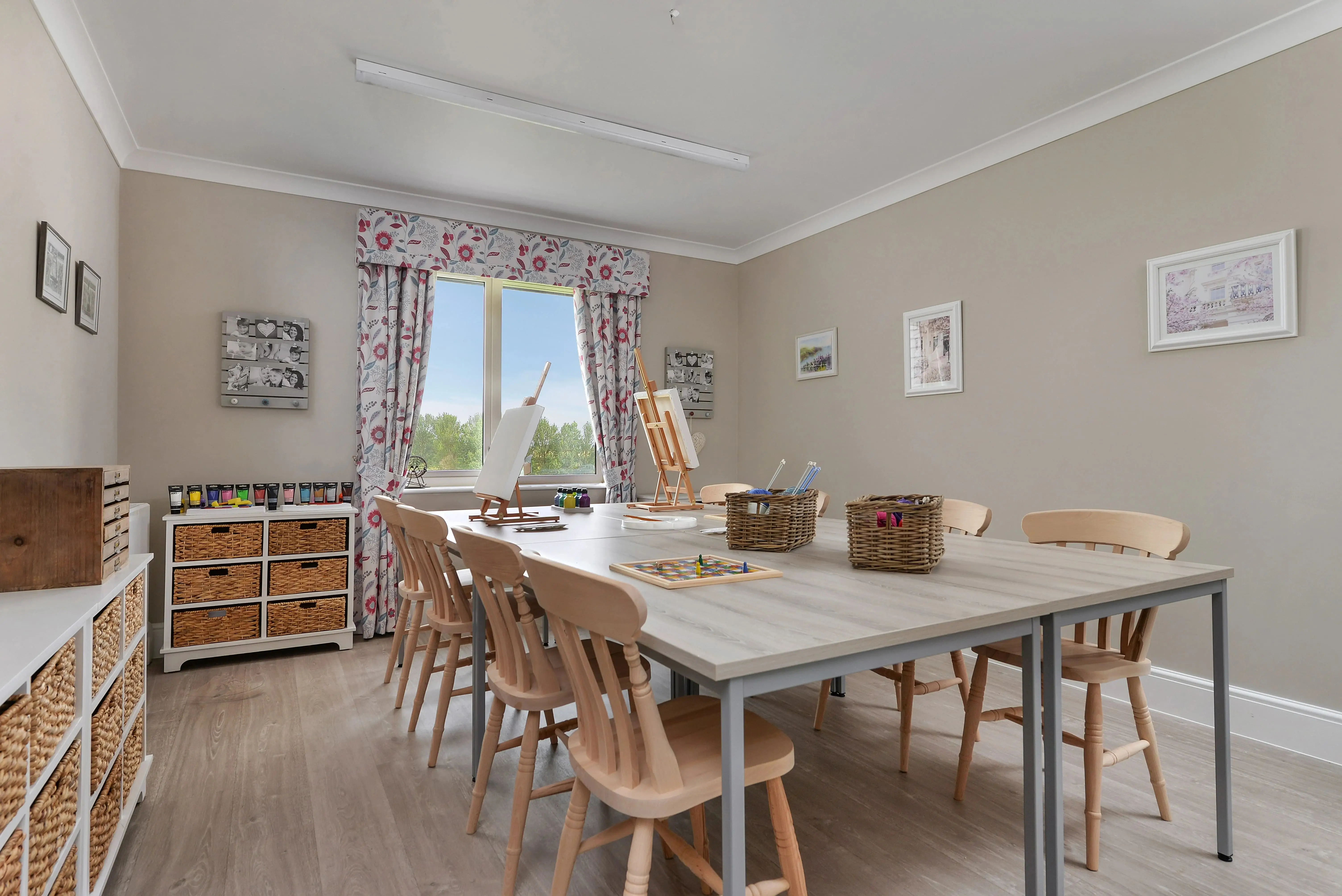 A spacious and organised craftroom with a large table, chairs, storage shelves filled with jars of paint, and an easel positioned by the window.