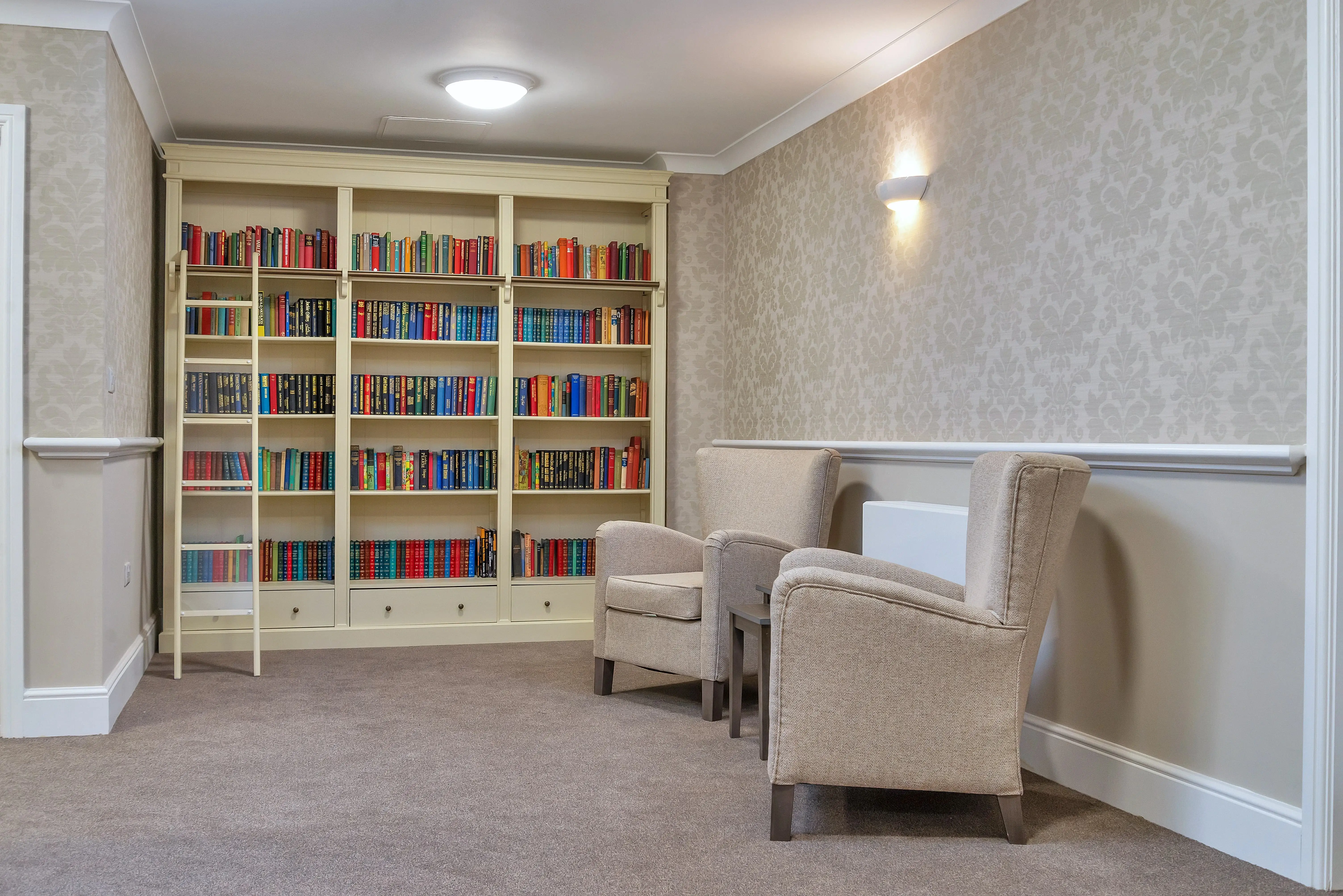 A cosy library with two beige armchairs facing a built-in bookshelf filled with colourful books